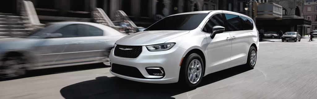 White Chrysler Pacifica minivan driving through a city street in an urban setting.